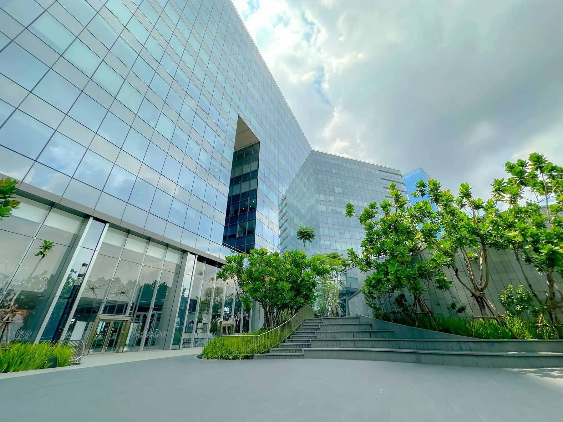 Contemporary glass office and mixed-use complex with a landscaped plaza and stepped terrace under a partly cloudy sky